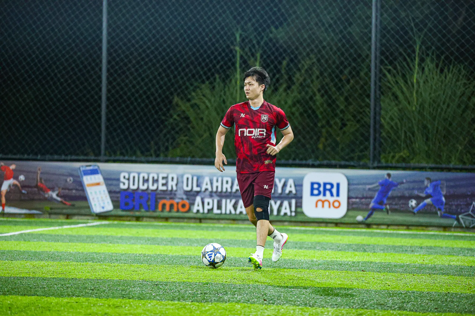 Pemain berjersey merah NOIR membawa bola di lapangan sintetis pada malam hari