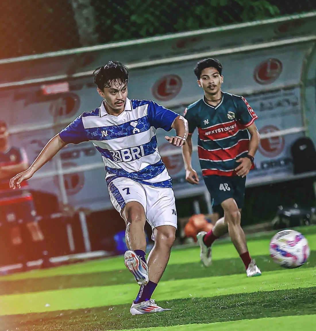 Pemain berjersey biru-putih bergaris NOIR menendang bola saat pertandingan malam
