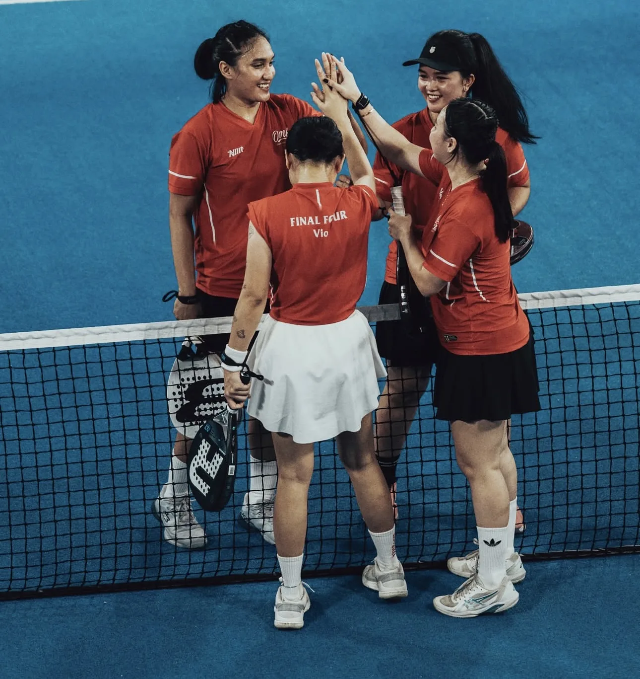 Pemain padel berjersey merah NOIR saling bertos di dekat net lapangan biru