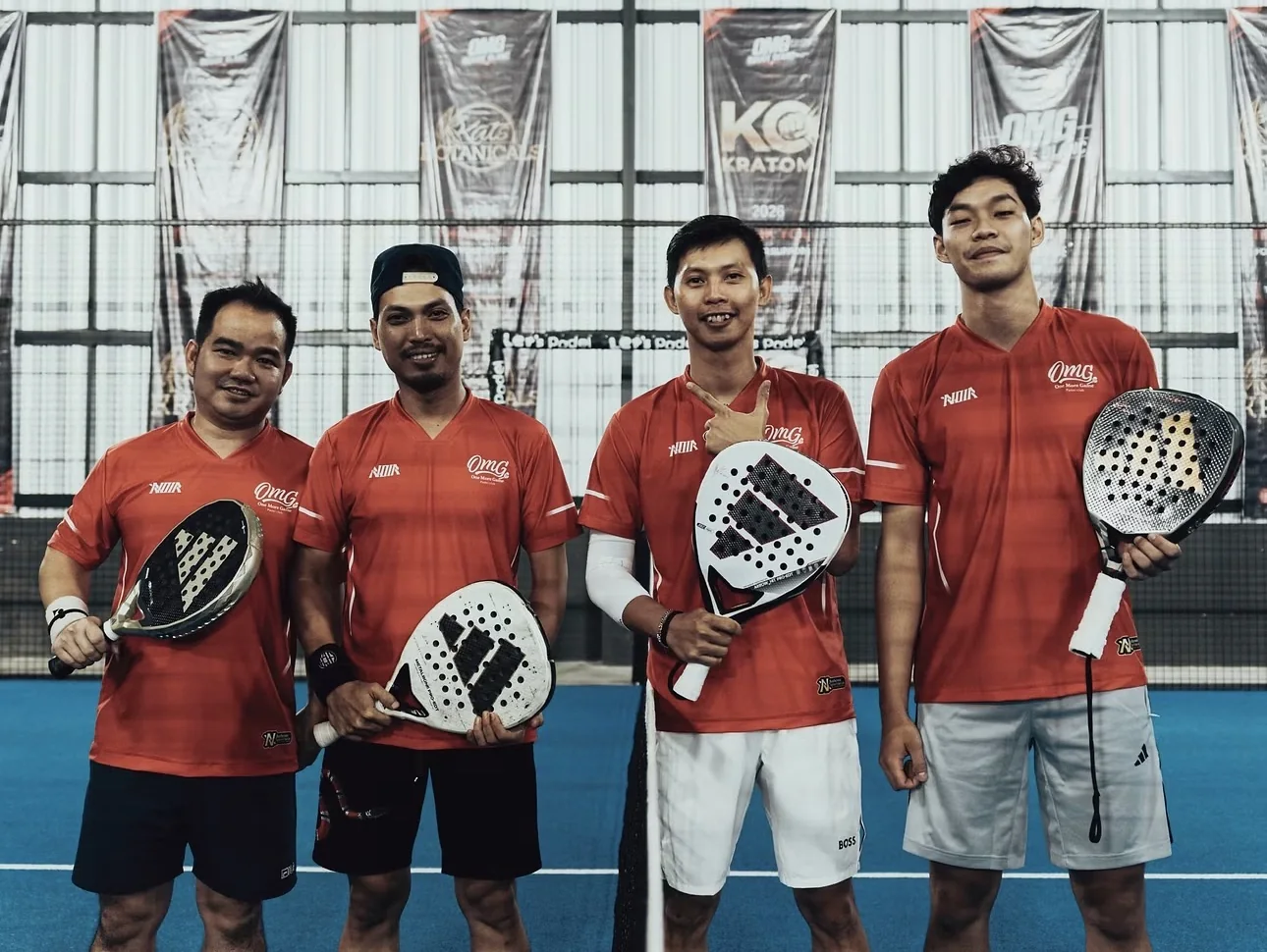 Empat pemain padel berjersey oranye NOIR memegang raket di lapangan biru, berdiri di dekat net
